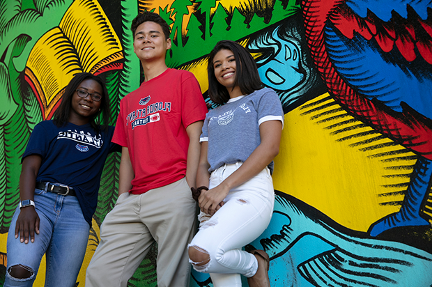 A male student and two female student lean against a wall covered with art